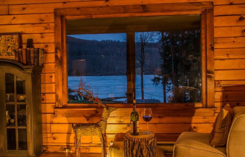 A warm ambiance is created by the wooden walls, while a large window frames a serene view of the frozen lake and surrounding trees. A decorative reindeer figurine sits beside a small wooden table with a wine glass and bottle.
