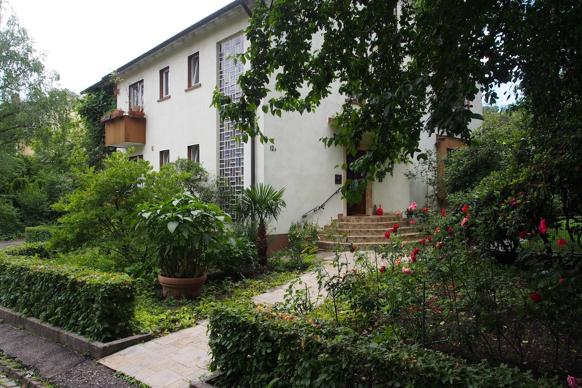 The residence is surrounded by lush greenery, featuring a neatly landscaped garden with a variety of plants and flowers. A stone pathway leads to the entrance, which is framed by steps and has multiple windows allowing natural light to fill the space.