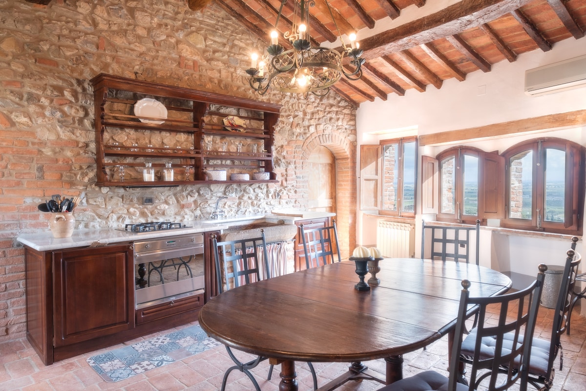 The kitchen showcases a blend of rustic and modern elements, featuring exposed stone walls and wooden beams. A round wooden table with six chairs is set, while a well-equipped kitchen area includes a gas stove, oven, and rustic shelving displaying dishware. Natural light illuminates the space.