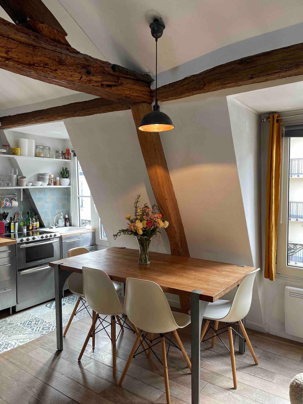 A dining area features a wooden table surrounded by four white chairs. An arrangement of flowers is placed in a glass vase at the center of the table. Exposed wooden beams ascend towards the ceiling, with a pendant light illuminating the space. A kitchen area is visible in the background.