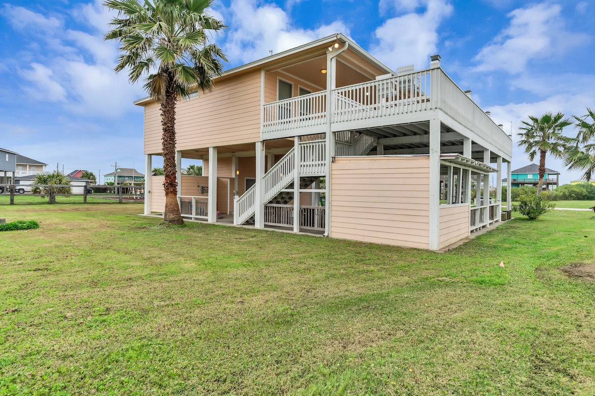 The exterior of a light-colored house is displayed, elevated on stilts with a spacious deck. Lush green grass surrounds the property, and palm trees offer a tropical feel. A staircase leads up to the deck, providing access to outdoor living spaces.