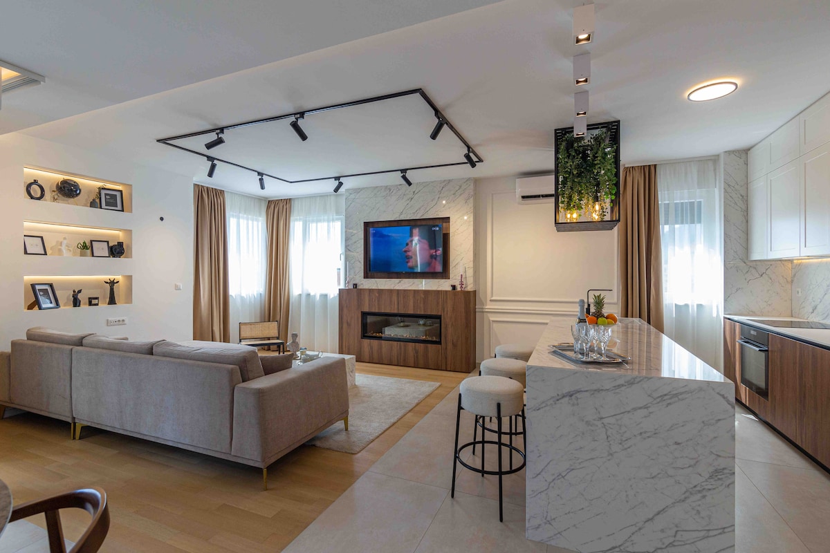 An open living area features a light-colored sofa arranged in front of a sleek entertainment unit with a television. Natural light enters through large windows, complemented by soft curtains. A breakfast bar with stools is visible, while decorative shelves showcase curated items.