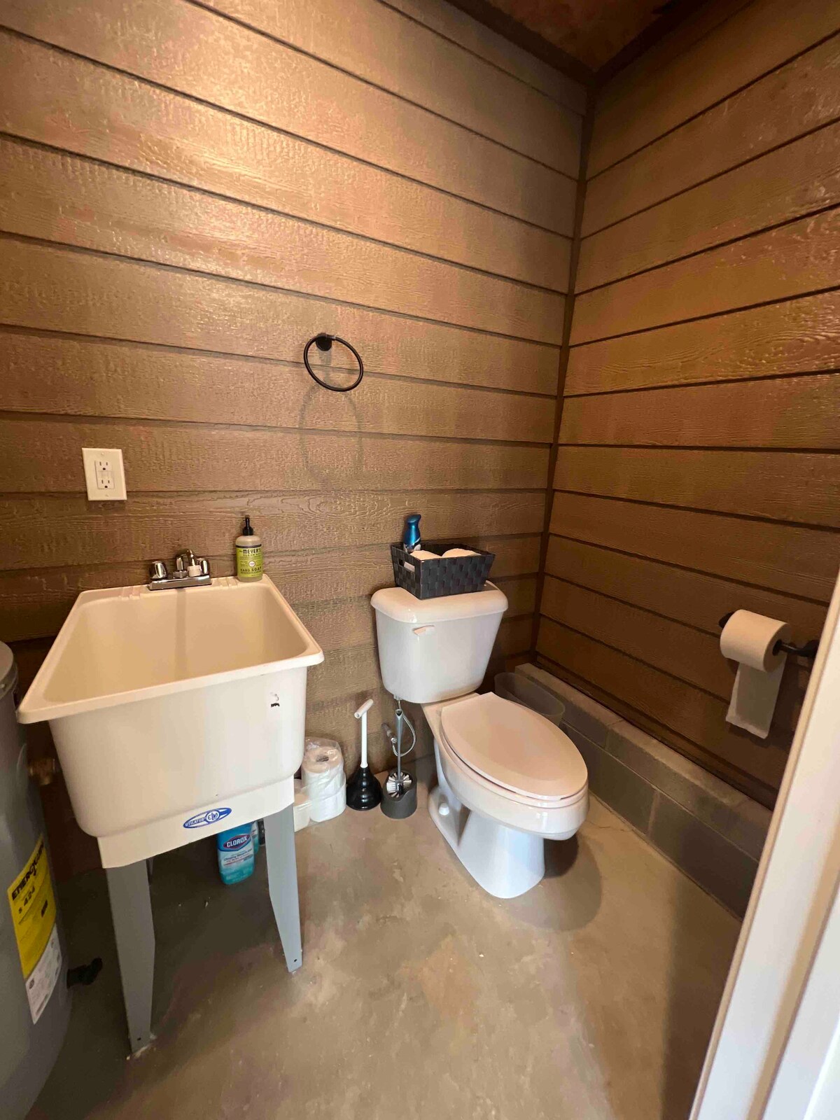A modern bathroom space features a compact sink situated beside a toilet. The walls are lined with warm wood panels, providing a cozy feel. Essential toiletries are arranged neatly, with a roll of toilet paper easily accessible. A concrete floor completes the functional design.