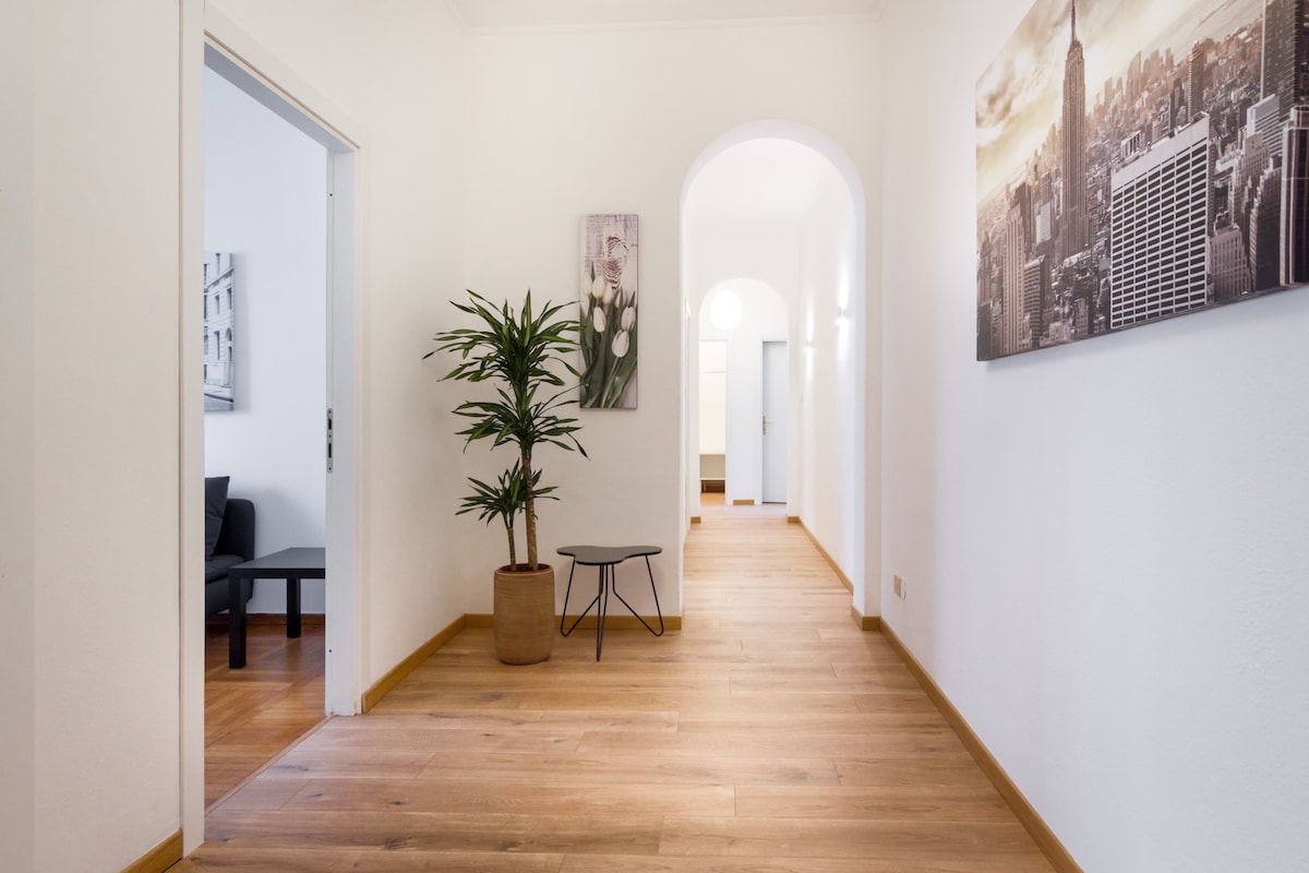 A bright hallway features light wooden flooring and white walls, creating an airy feel. Potted plants add a touch of greenery, while framed images of cityscapes adorn the walls. Doorways lead to additional rooms, enhancing the apartment's spacious layout.