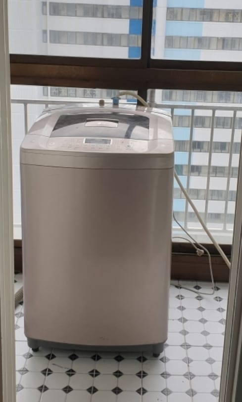 A washing machine is positioned on a balcony, featuring a sleek design with control buttons on the top. The tiled floor beneath is white with a geometric pattern. A view of a tall building is visible through the balcony railing.