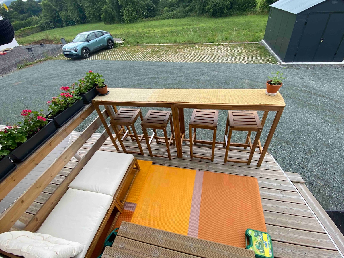 An outdoor seating area is presented with wooden bar stools arranged around a tall table. Potted flowers add a touch of greenery, while a cozy seating area is positioned beside an orange rug. A view of the surrounding grass and gravel is visible in the background.