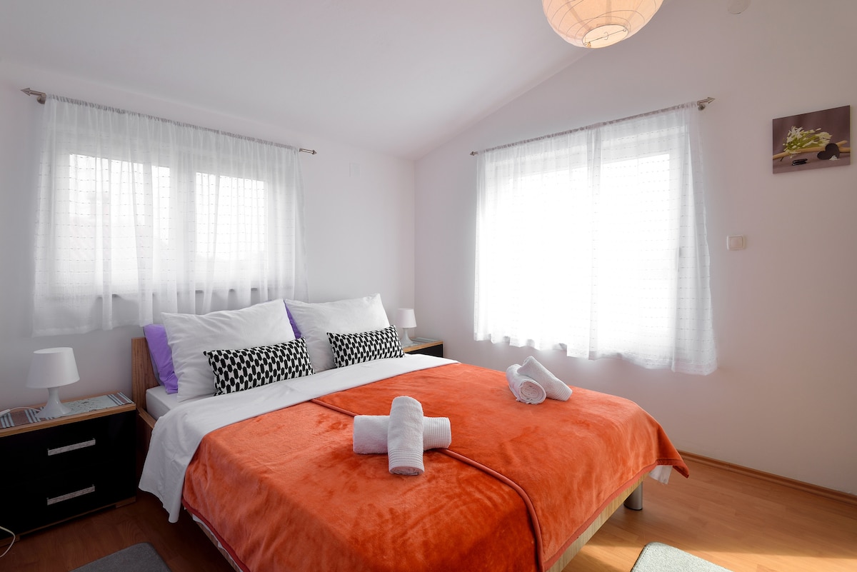 A well-lit bedroom features a spacious bed adorned with vibrant orange bedding and accented by patterned pillows. Two bedside lamps provide gentle illumination, and large windows are dressed with sheer curtains, allowing natural light to brighten the room.
