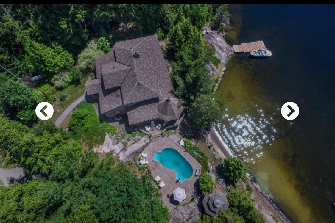 An aerial view of a spacious cottage surrounded by lush greenery and waterfront. A private in-ground pool is visible in the center, complemented by a gazebo nearby. A dock extends into the water, enhancing the natural setting.