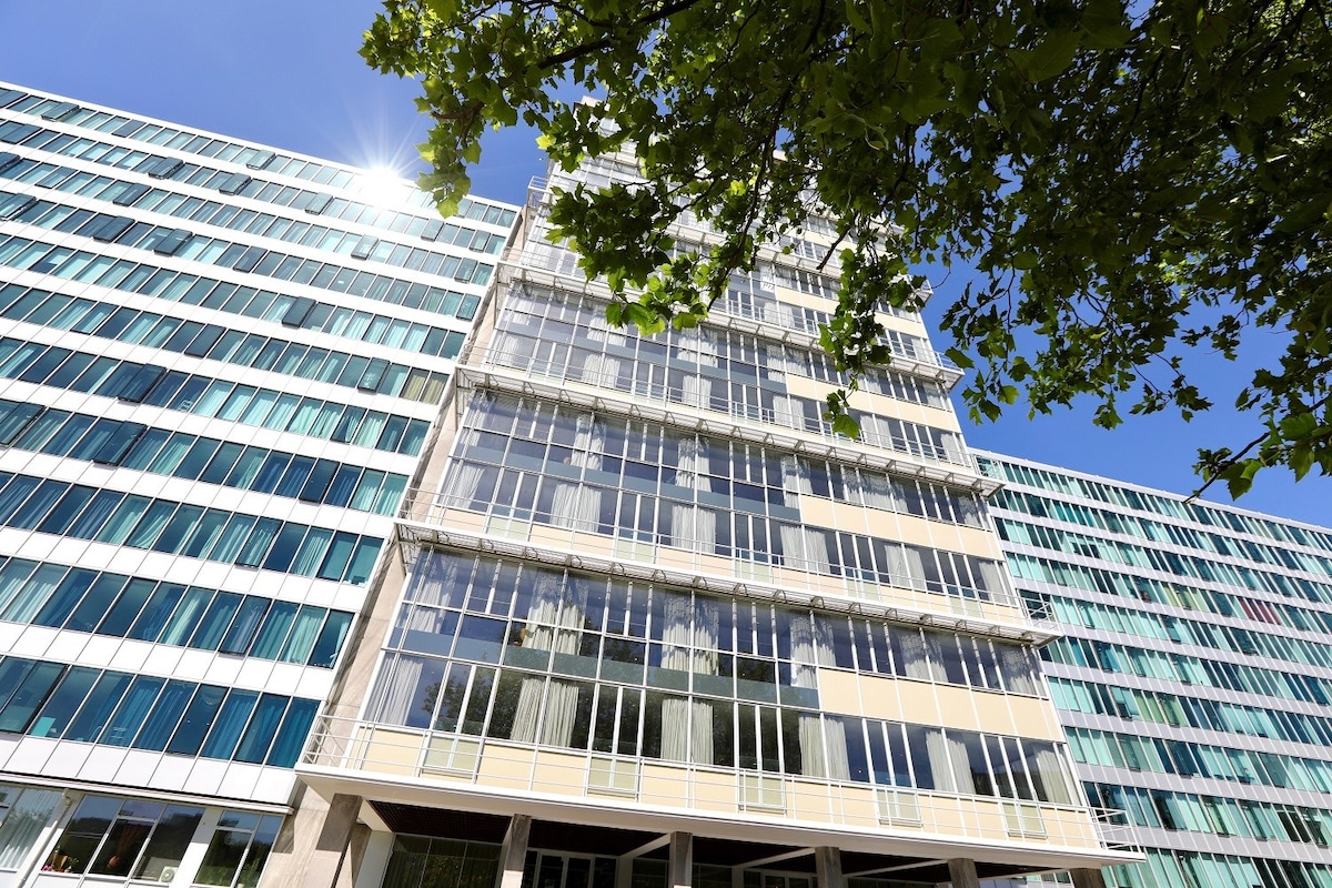 A modern building showcases a façade of glass and contemporary architecture. The structure features multiple stories with large windows reflecting natural light. Lush greenery is visible in the foreground, complementing the vibrant atmosphere.