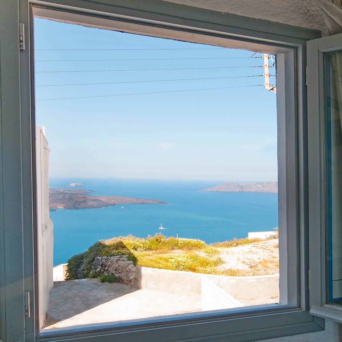 A view from a window reveals expansive blue waters and distant islands. The scene showcases the natural beauty of the coastline, framed by the light-colored window trim. Bright flowers can be seen in the foreground, adding a touch of color to the serene landscape.