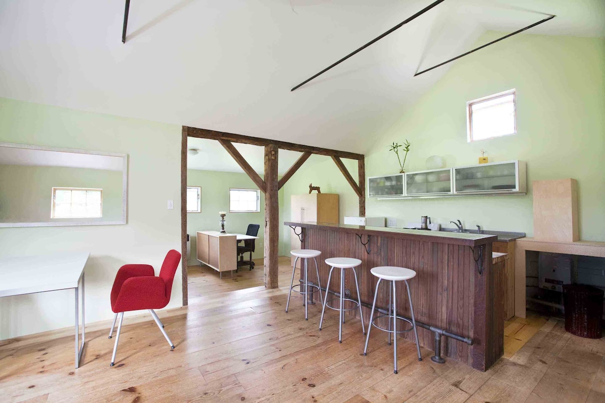 An open kitchen space features light green walls and natural wood flooring. A kitchen island with seating for three is present, alongside a sleek dining table with a red chair. Clear cabinetry showcases minimalist design, while exposed wooden beams add character to the room.