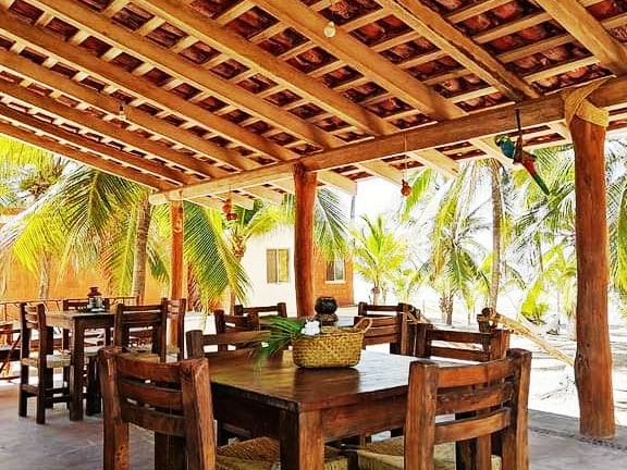An open-air dining area features rustic wooden tables accompanied by sturdy chairs, set beneath a patterned wooden ceiling. Lush palm trees provide natural greenery, while decorative items enhance the inviting space, creating a relaxed venue for meals or gatherings.