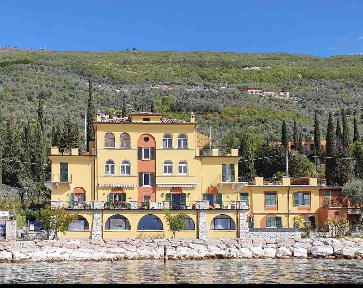 A vibrant building stands along the waterfront, showcasing a blend of yellow and orange tones. Multiple large arched windows are present, providing views of the surrounding landscape. Towering cypress trees and lush greenery frame the structure, enhancing its proximity to the serene shoreline.