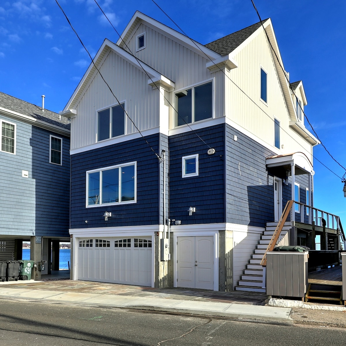 The exterior of a newly constructed home features a combination of blue and white siding. Large windows are present on multiple levels, allowing for ample natural light. A staircase leads to the entrance, while parking is available beneath the house.