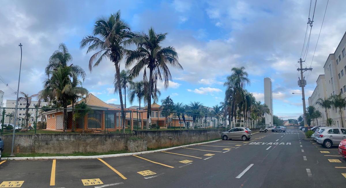A spacious outdoor area is lined with palm trees, providing a tropical ambiance. The setting features a well-maintained building in the background, and a parking lot with marked spaces is visible on the side of the road, indicating easy accessibility.