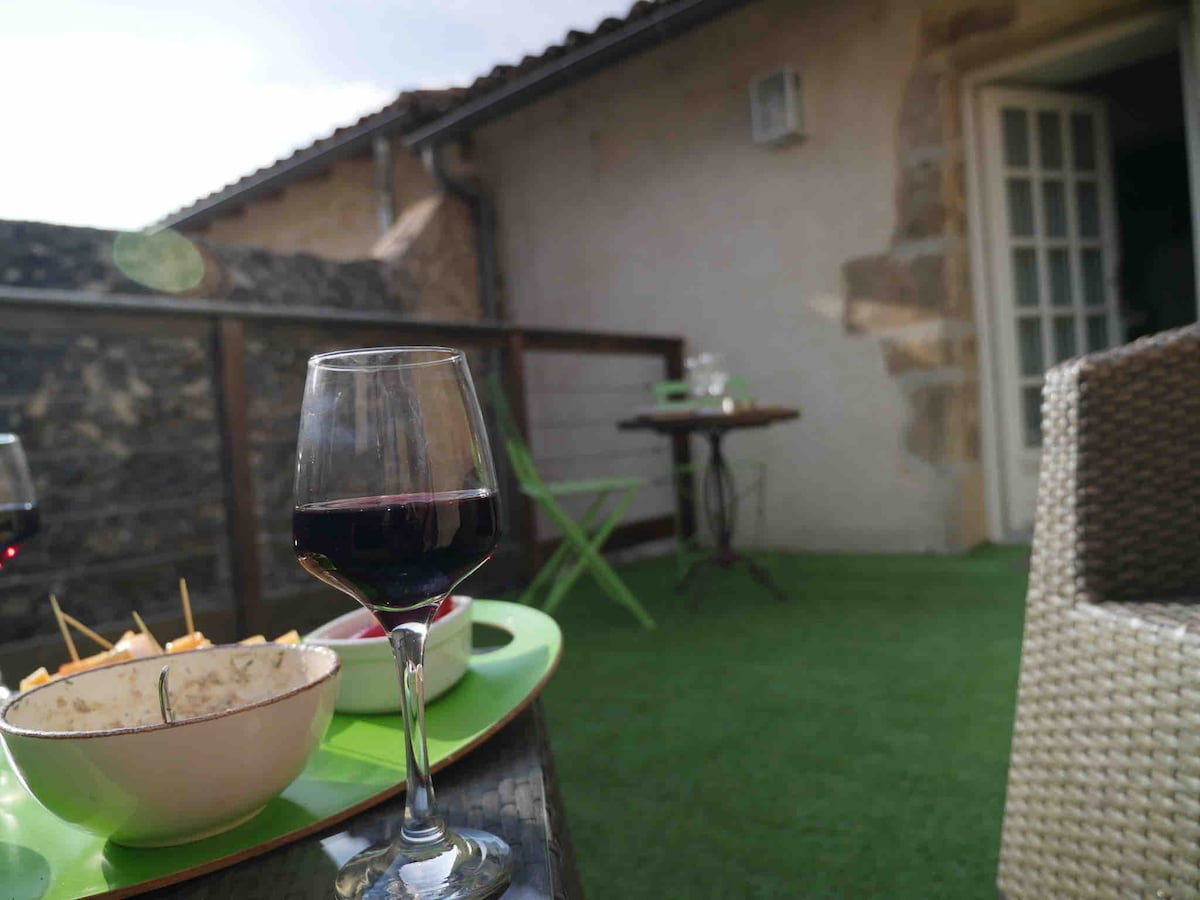 A terrace is visible with green artificial grass, featuring a small table and chairs. A glass of red wine rests on a tray alongside a bowl, partly filled with food. Natural light filters in from a nearby open door, creating a serene outdoor setting.