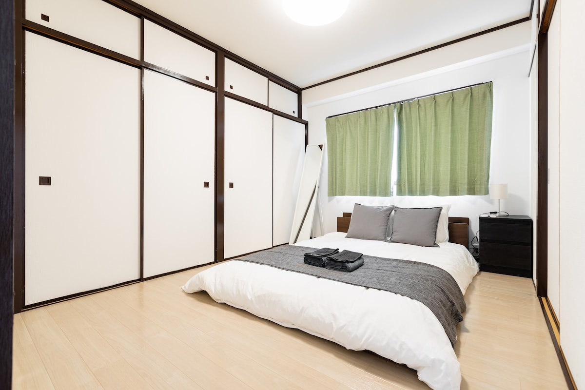 A spacious sleeping area features a low-profile bed with a neatly arranged blanket and towels. Large windows are dressed in green curtains, allowing natural light to fill the room. Built-in closets offer ample storage space, while minimalistic furniture complements the simple aesthetic.