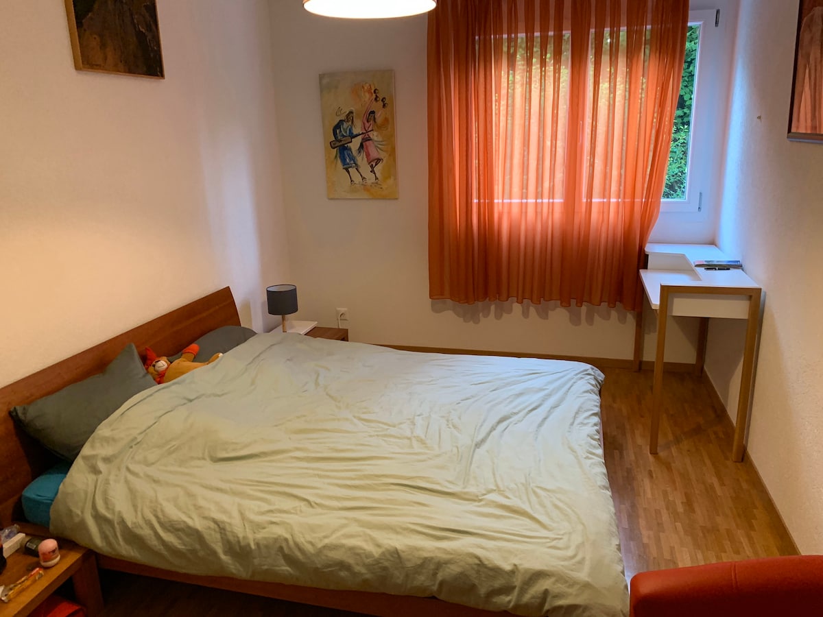 A sleeping area features a spacious bed dressed in light bedding, complemented by decorative pillows. Natural light filters through the orange curtains, illuminating the warm wood flooring. A small desk and a lamp are visible beside the bed, providing functional elements to the space.