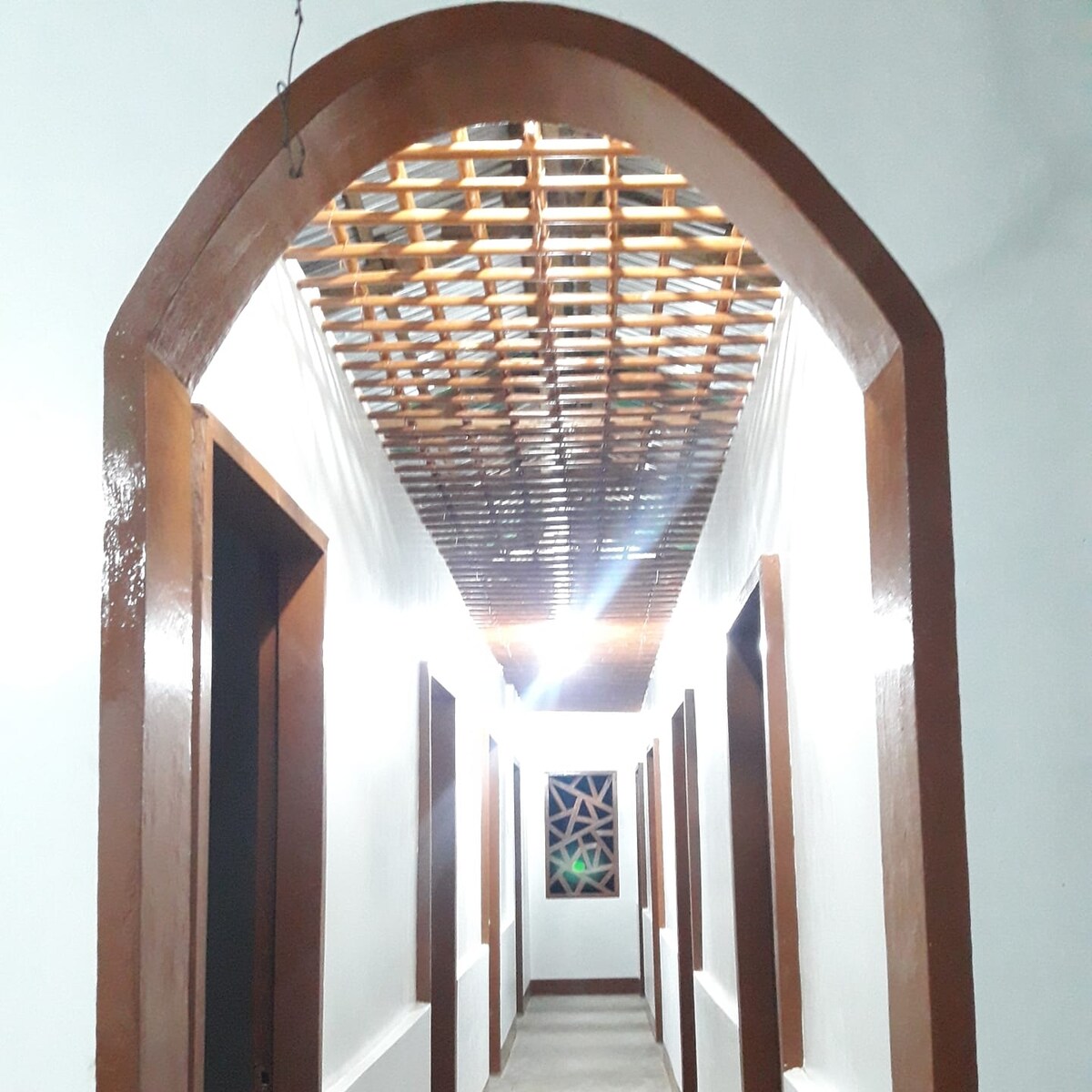 A well-lit hallway features an arched ceiling made of woven materials, creating a natural aesthetic. The walls are painted white, enhancing brightness, while wooden doors line either side, leading to private rooms. A decorative window is visible at the end of the corridor, adding charm.