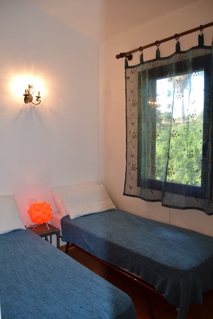 Ბინის მეორე ოთახი: ორი კომფორტული საწოლი, მოდის აბატჟური და ფანჯარა, რომელიც ხმელთაშუა ზღვის მცენარეულობაზეა.