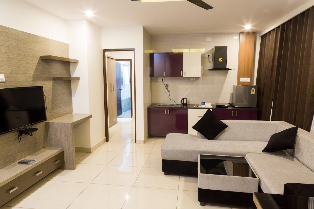 A modern living space features a sleek gray and purple kitchenette with essential appliances such as a kettle. A plush sectional sofa with black accent pillows provides seating. A wall-mounted flat-screen TV is visible, alongside minimalist shelving units, creating a spacious and functional environment.