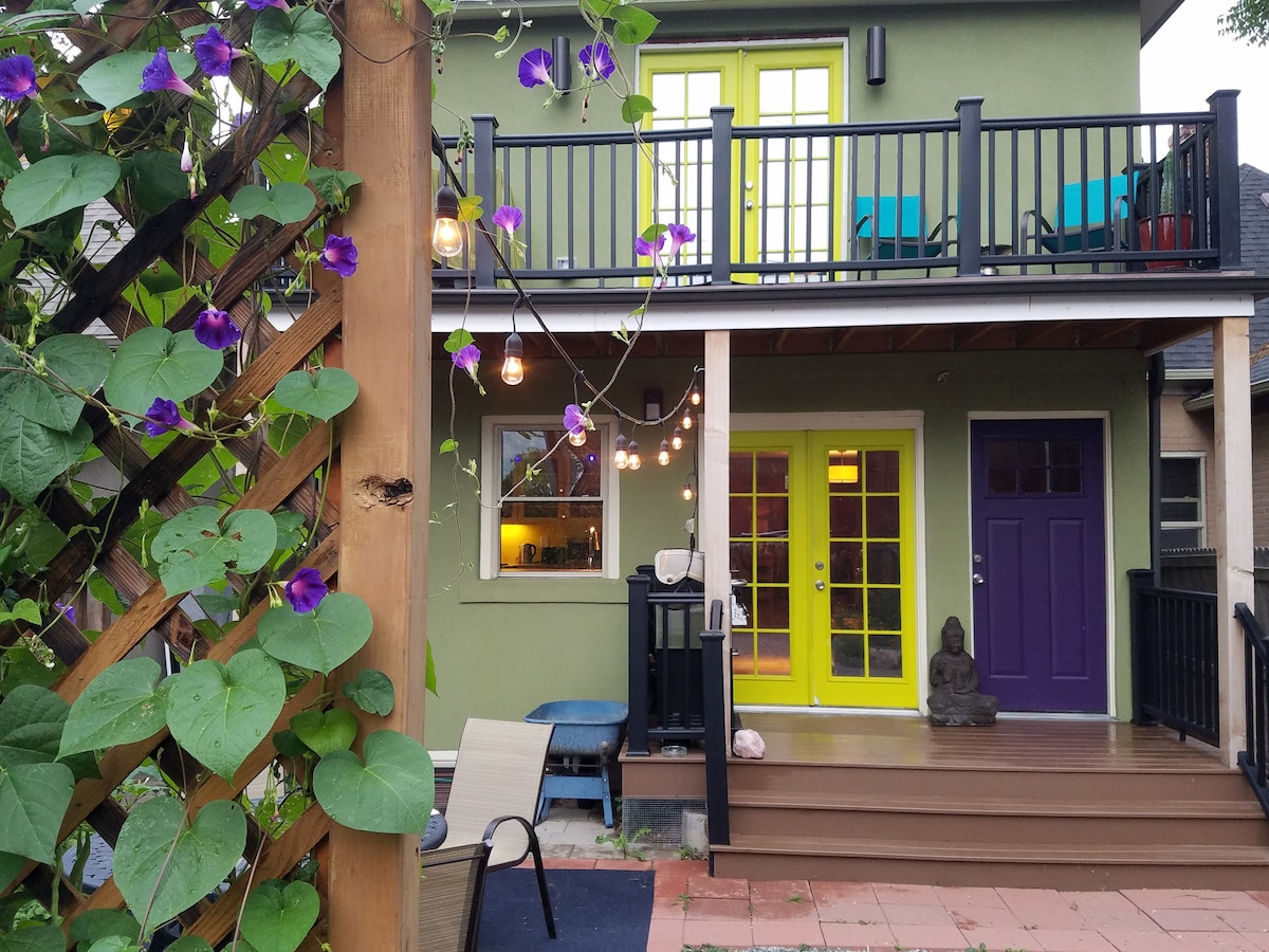 A contemporary home features a vibrant green exterior complemented by purple and yellow doors. A spacious deck is visible above, adorned with colorful chairs. Climbing plants and string lights enhance the outdoor space, with a peaceful statue nestled nearby, creating a welcoming entrance.