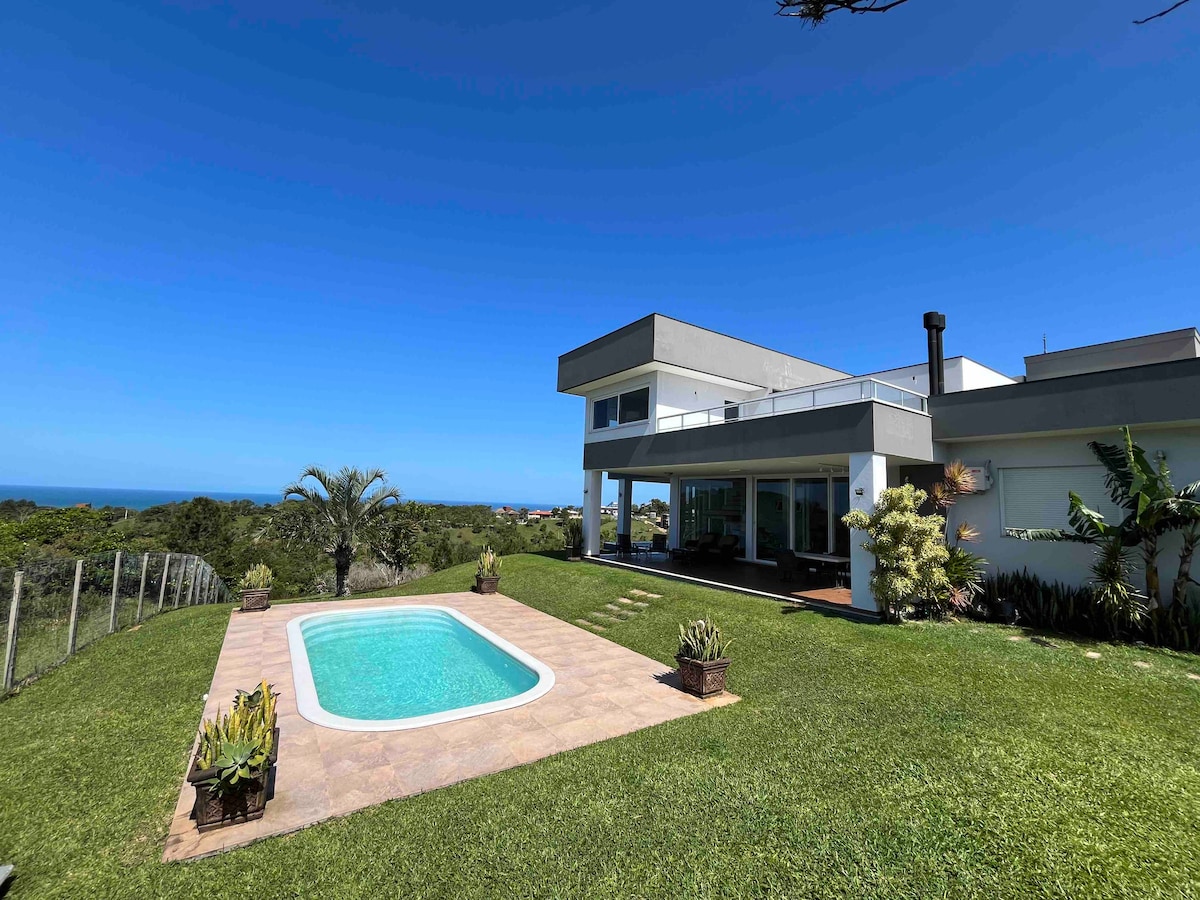 A modern house is set on a hillside, featuring a private swimming pool surrounded by a neatly landscaped garden. Expansive windows offer panoramic views of the ocean and surrounding greenery. The patio area includes tropical plants and a well-maintained lawn.