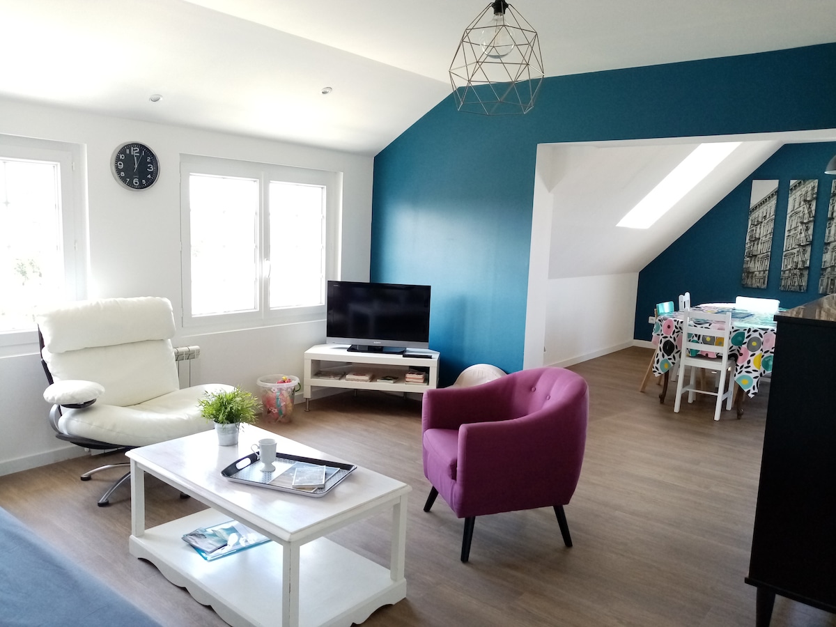 A light-filled living area features a white coffee table and a comfortable armchair in soft purple. A modern television rests on a low wooden cabinet, complemented by a wall clock above. Nearby, a dining area showcases a table adorned with colorful decor against a contrasting blue wall.