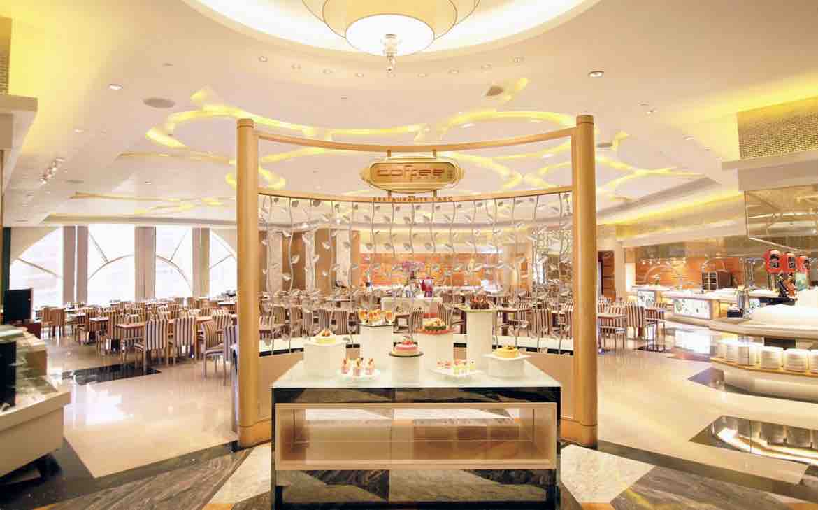 An elegant dining area is showcased with modern architecture. Stylish wooden chairs are arranged around tables, while a dessert display is featured at the center. Soft, warm lighting enhances the ambiance, and large windows allow natural light to fill the space.