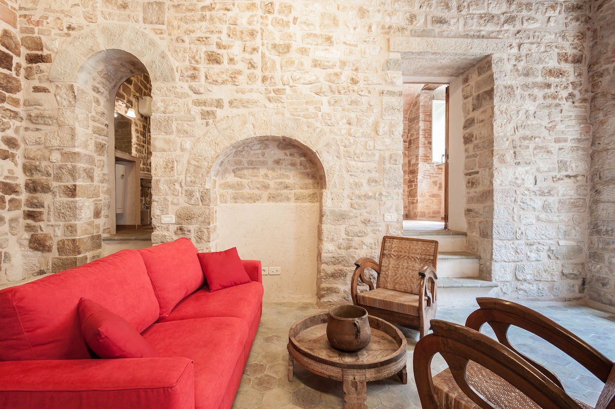 A living area features exposed stone walls with medieval arches, creating a historic ambiance. A red sofa with cushions is positioned next to a round wooden coffee table. Two wooden chairs provide additional seating, complementing the rustic charm of the space.