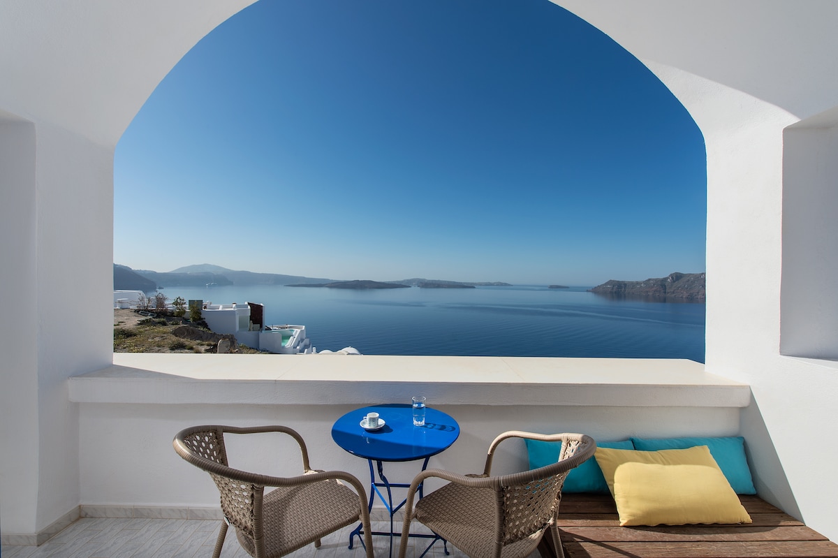 A private balcony features two woven chairs and a small blue table, inviting relaxation. The expansive view showcases the tranquil Aegean Sea, framed by distant hills and the clear blue sky, creating a serene atmosphere. A yellow cushion adds a touch of color to the setting.