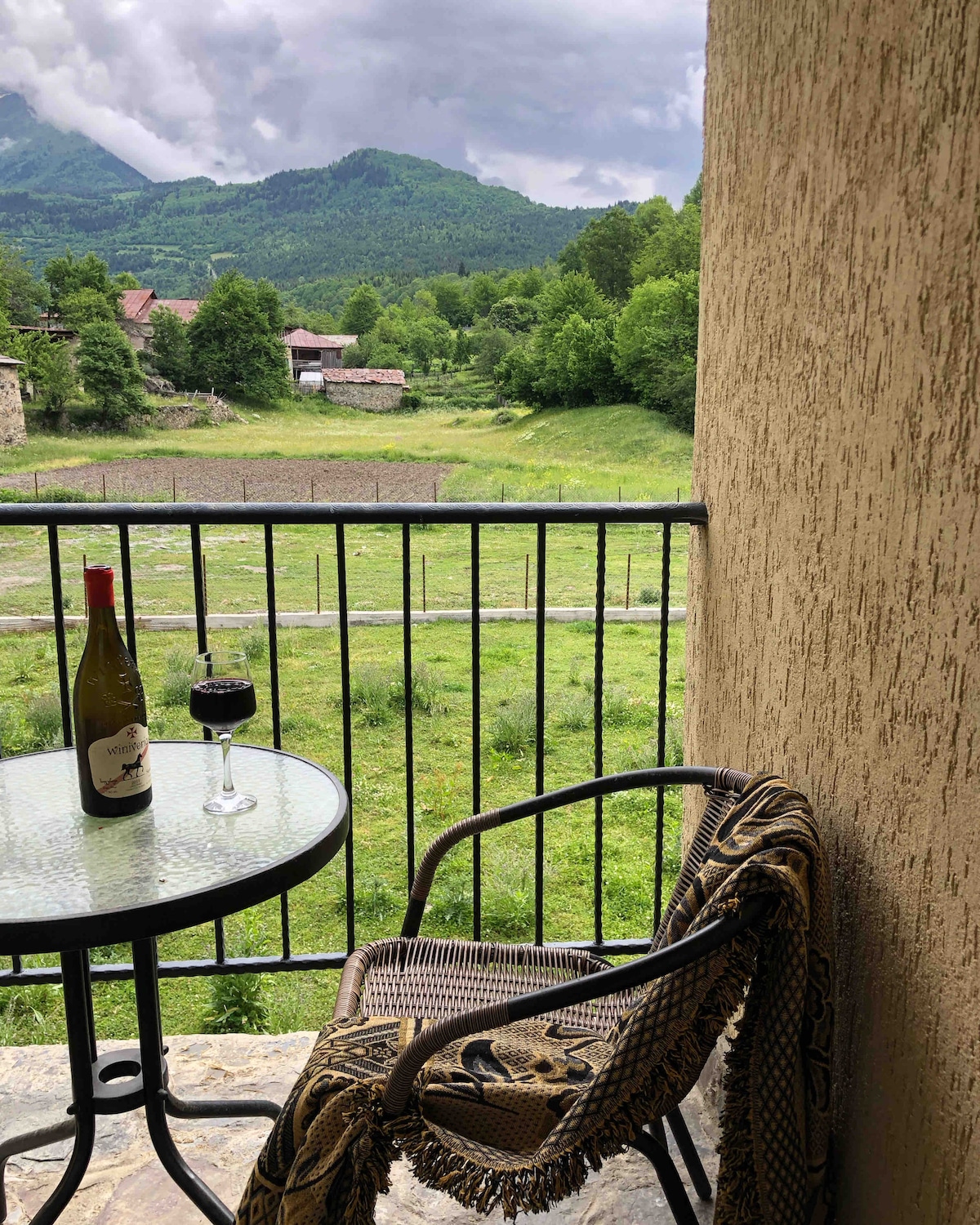A small balcony is showcased with a round table set for two. A bottle of wine and a glass are placed on the table, while a woven chair, draped with a cozy blanket, offers a view of the green valley and distant mountains.
