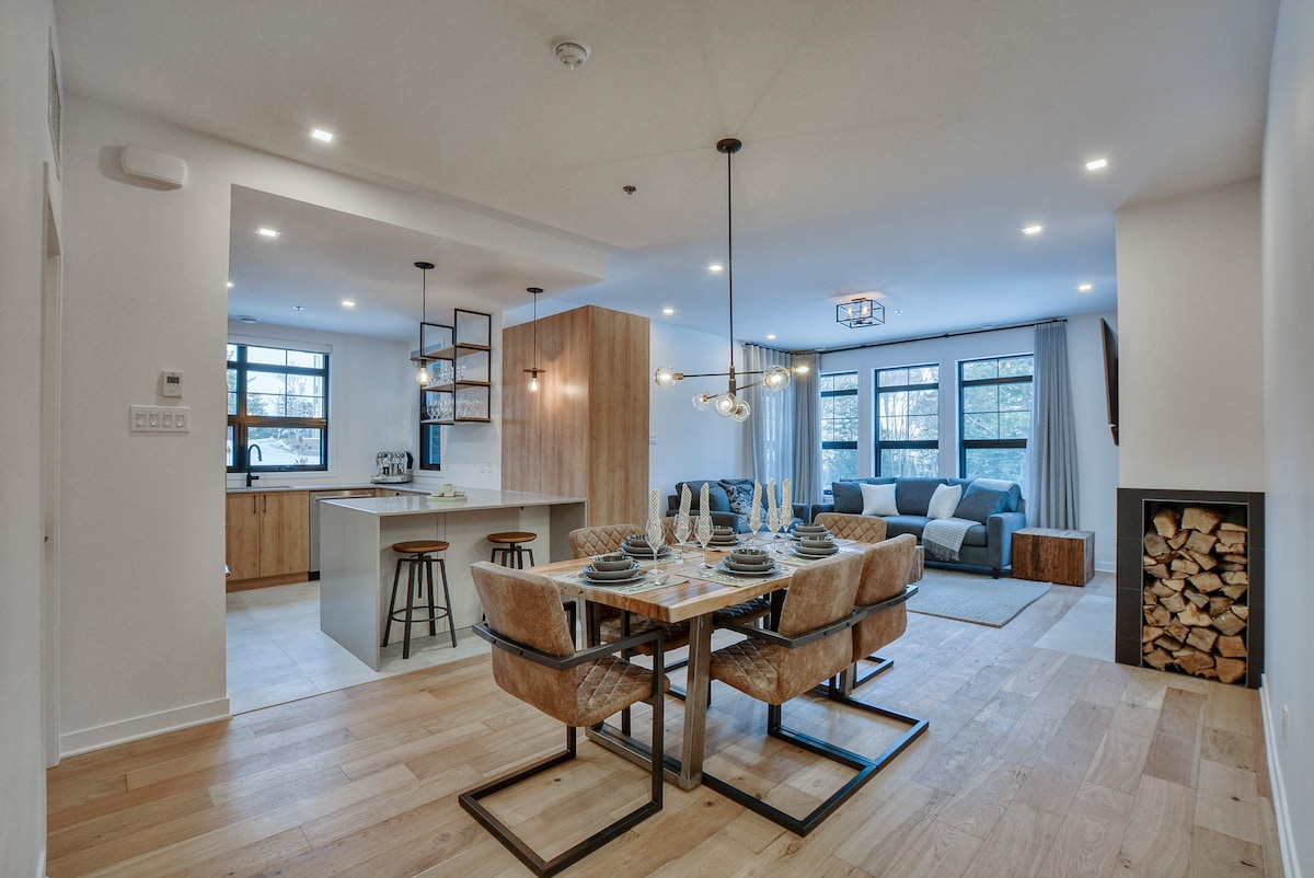 An open-concept living and dining area displays a modern design, featuring a dining table set for four alongside comfortable seating. Large windows allow natural light to fill the space, while a stylish kitchen with bar seating complements the inviting atmosphere.