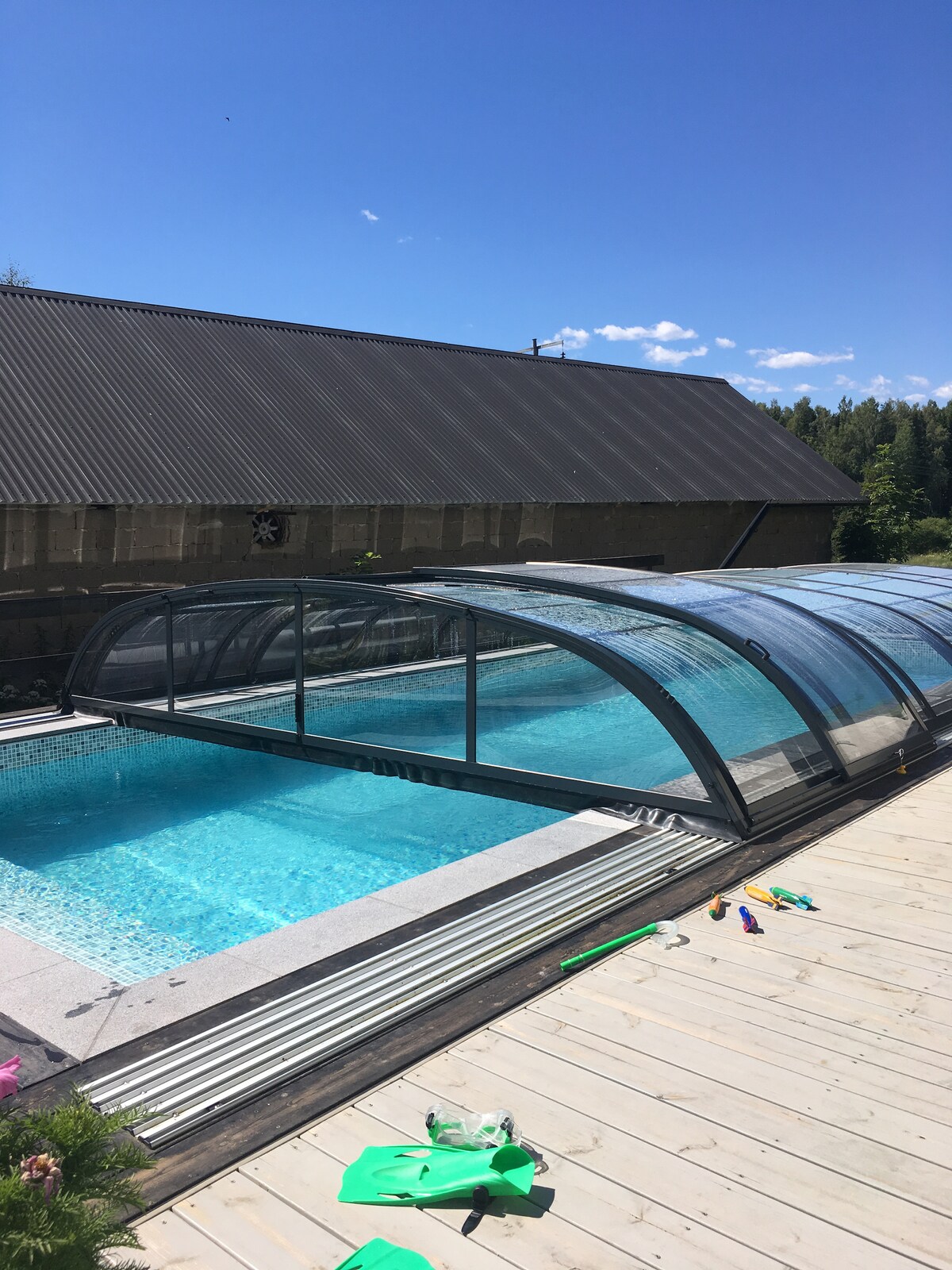 An outdoor pool is enclosed by a transparent cover, allowing sunlight to reflect off the water's surface. Surrounding decking provides space for relaxation, while a sturdy building with a metal roof sits in the background, complemented by clear blue skies.
