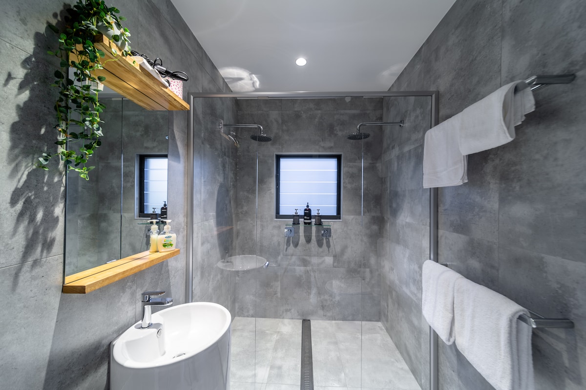 A modern bathroom is highlighted by gray tiled walls and floors. A glass walk-in shower features dual showerheads, and white towels hang neatly on silver racks. A round sink is positioned below a wooden shelf adorned with greenery, complemented by a mirrored wall.