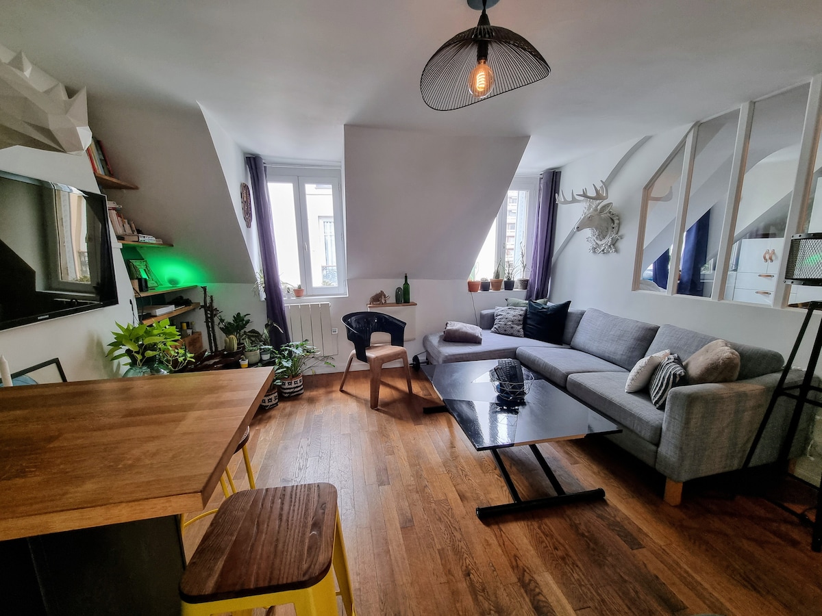 A bright living area is presented, featuring a comfortable grey couch and a stylish coffee table. Floor-to-ceiling windows allow natural light to fill the space, framed by decorative plants. A dining area is visible with modern chairs, complemented by unique wall décor.