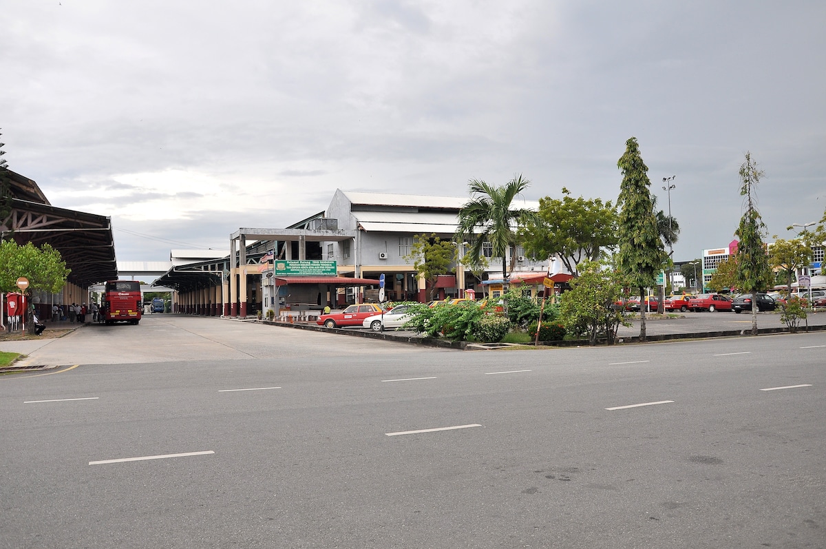 A view of Shahab Perdana Bus Terminal is displayed, showcasing a spacious area with parked buses and cars. Lush greenery is present along the sides, with palm trees and shrubs enhancing the landscape. The terminal building features a modern design, visible on the right side of the image.
