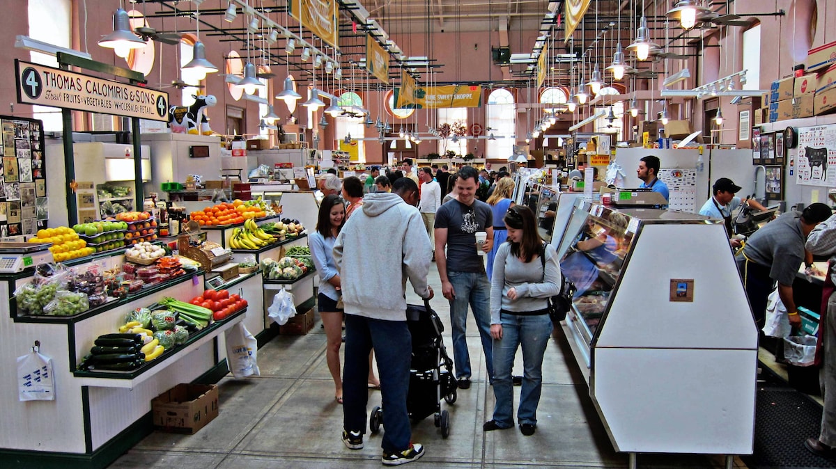 An indoor market bustling with activity. Colorful displays of fruits and vegetables fill each stall, while shoppers browse local goods. Bright overhead lights illuminate the space, enhancing the lively atmosphere. Various counters feature fresh produce, meats, and baked items, creating a vibrant shopping experience.