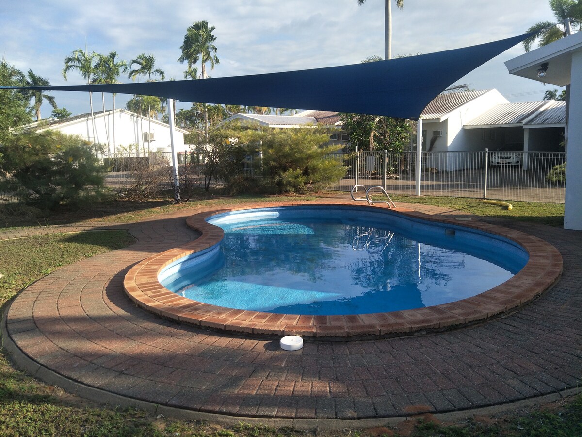 A swimming pool is surrounded by a circular brick patio, featuring a shaded area overhead. Lush greenery borders the pool area, offering a peaceful outdoor retreat. The pool's clear water reflects the sky, enhancing the overall tranquil setting.