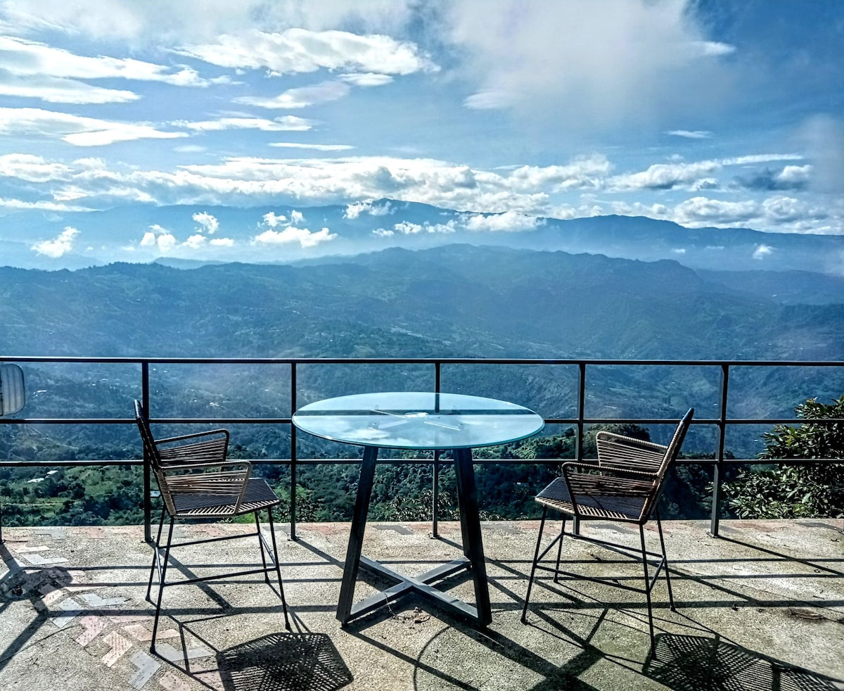 A transparent glass table is centered on a terrace, accompanied by two woven chairs. A railing offers unobstructed views of the mountainous landscape, with distant hills and a partly cloudy sky in the background.
