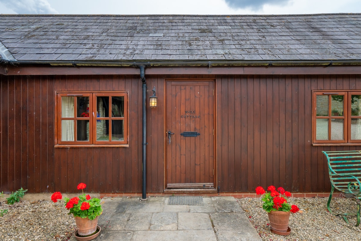The exterior of Well Cottage is composed of dark wood cladding, featuring a wooden door with a simple sign above. Two flower pots with vibrant red geraniums frame the entrance, and a green bench is positioned nearby on a stone pathway.