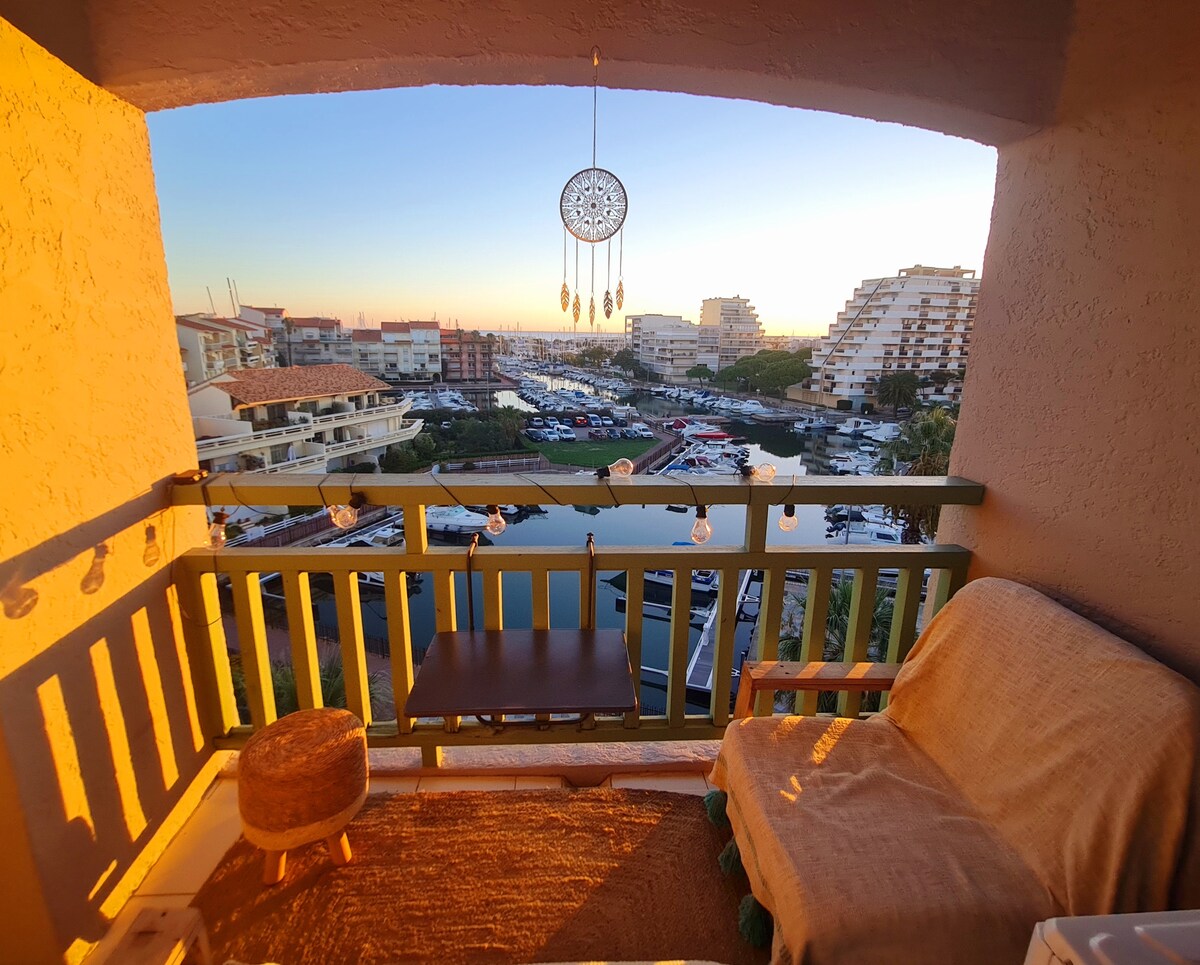 A cozy balcony is visible, featuring a small table and a comfortable seat. Strings of lights provide gentle illumination. The view includes a marina with boats and buildings in the background, while the sunset casts a warm glow over the scene.