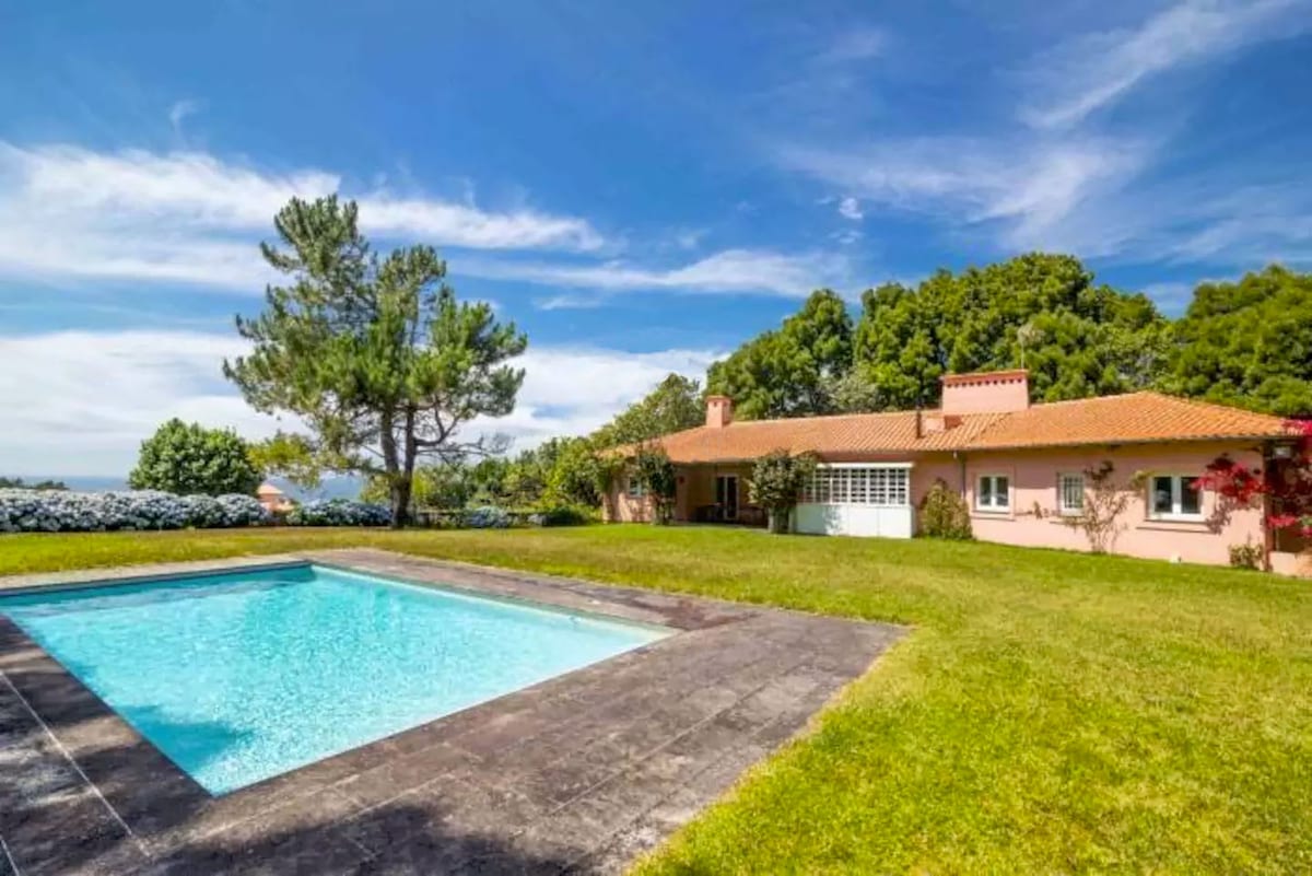 A spacious outdoor area features a private pool surrounded by manicured grass. The house, painted in soft pink tones, offers multiple windows and a red-tiled roof set against a backdrop of lush trees and a clear blue sky.