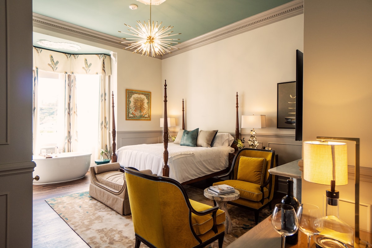 The guest suite features a four-poster bed dressed in soft linens. A freestanding soaking tub is positioned near large windows framed by patterned curtains. A cozy seating area includes two armchairs with a decorative rug beneath. Light fixtures add warmth to the space.