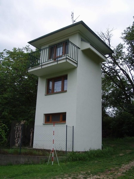 A two-story house is depicted, featuring a sleek exterior painted in light color with a flat roof. The balcony is accessed through large windows, while two smaller windows are situated below. Lush greenery surrounds the base, enhancing the house's unique structure.