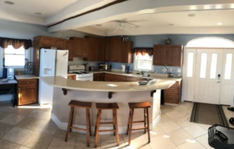 The kitchen features wooden cabinetry and a large island with two stools. A white refrigerator and appliances are visible, along with a window allowing natural light. The entryway is seen in the background, showcasing a front door and tiled floor.