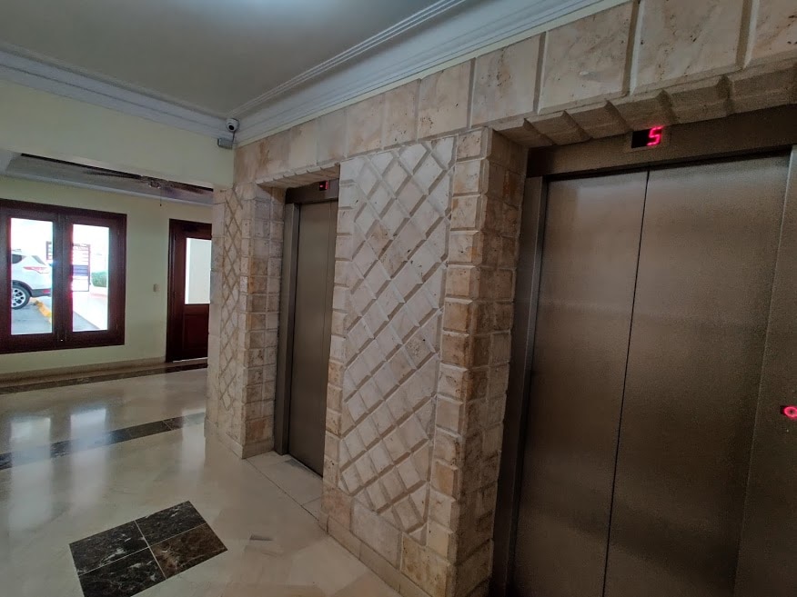 The image showcases two elevators located in a well-lit lobby area. The walls are adorned with textured stone, and the floor features an elegant marble design. Large windows allow natural light to fill the space, enhancing its welcoming appearance.