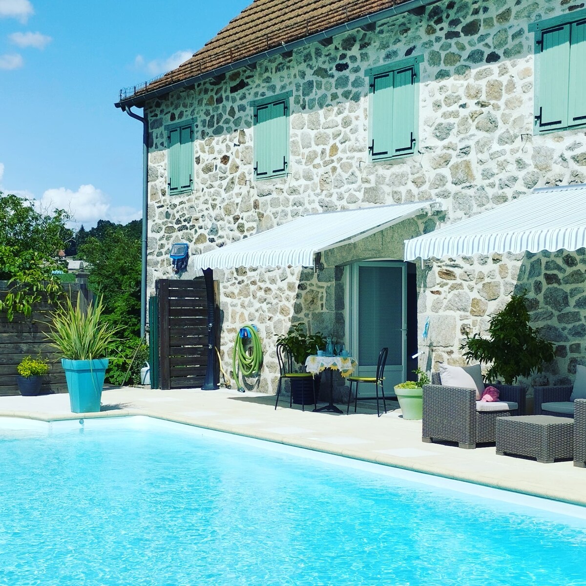 A stone building features light blue shutters and a covered terrace with dining furniture, situated beside an inviting swimming pool. Lush greenery and potted plants enhance the serene outdoor setting, while a clear sky completes the peaceful atmosphere.