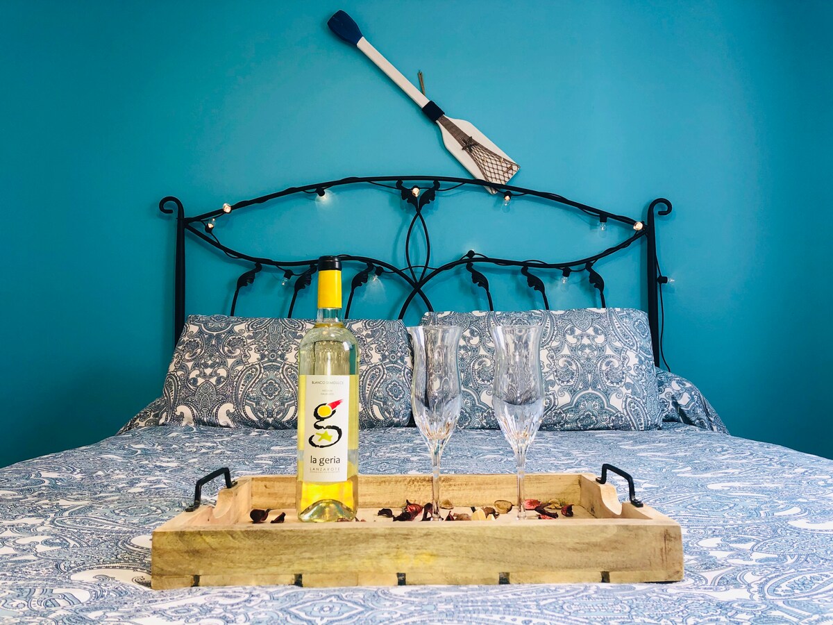 A spacious bed features a decorative blue wall and patterned bedding. A wooden tray holds a bottle of white wine and two glasses, set against the backdrop of an artistic oar mounted on the wall. Soft lighting complements the cozy atmosphere.