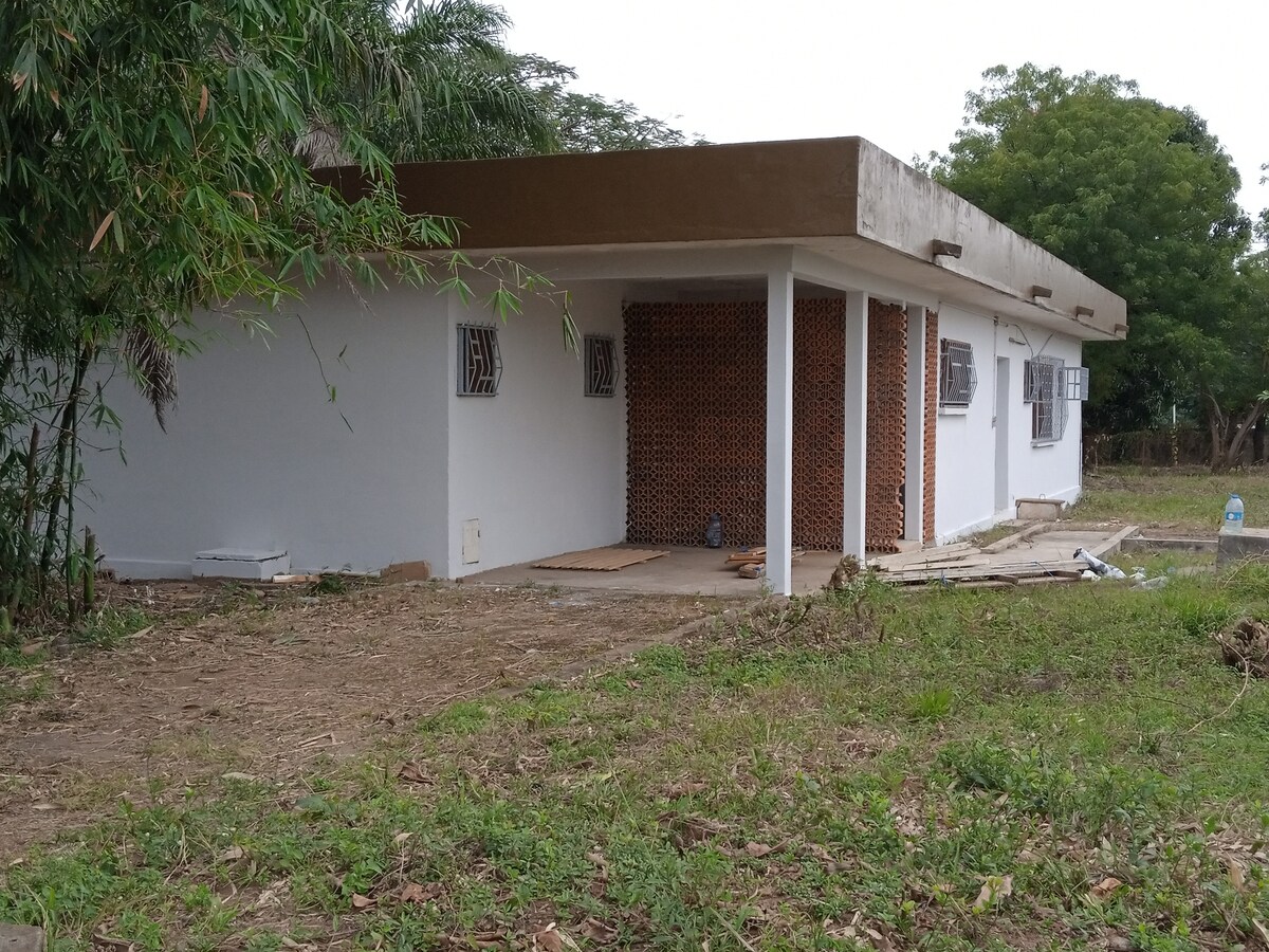 A simple exterior view of the villa, showcasing white walls with a brown overhang. The building features several windows and a textured brick section. Surrounding greenery and untended land create a natural environment, enhancing the setting's tranquility.